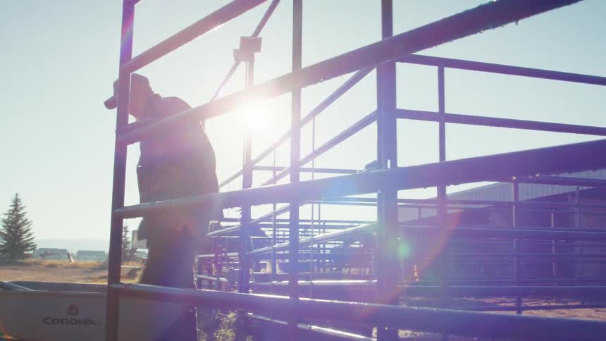 A farmer with a white beard opens a metal gate on a sunny ranch before pushing a wheelbarrow into the corral