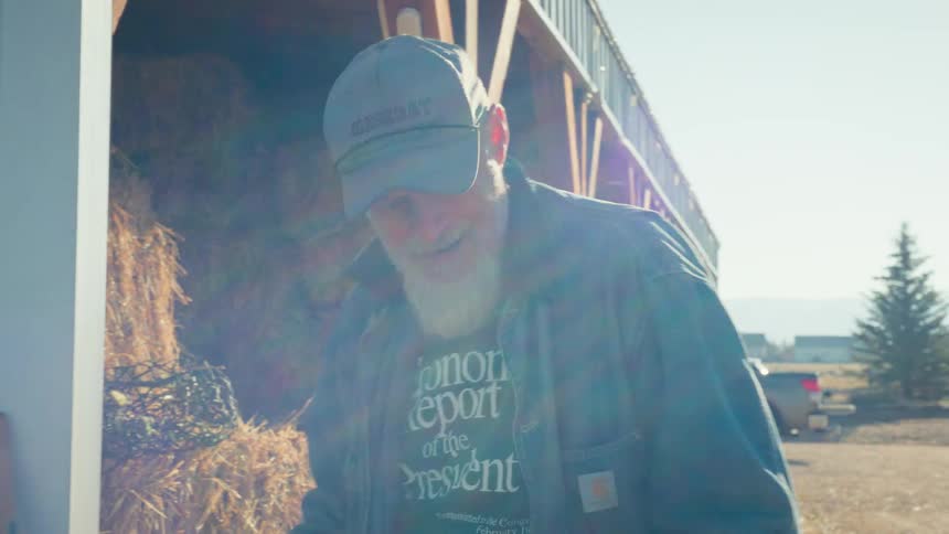 A hardworking older farmer with a long white beard smiles contentedly while working by the hay barn on a bright, sunny day
