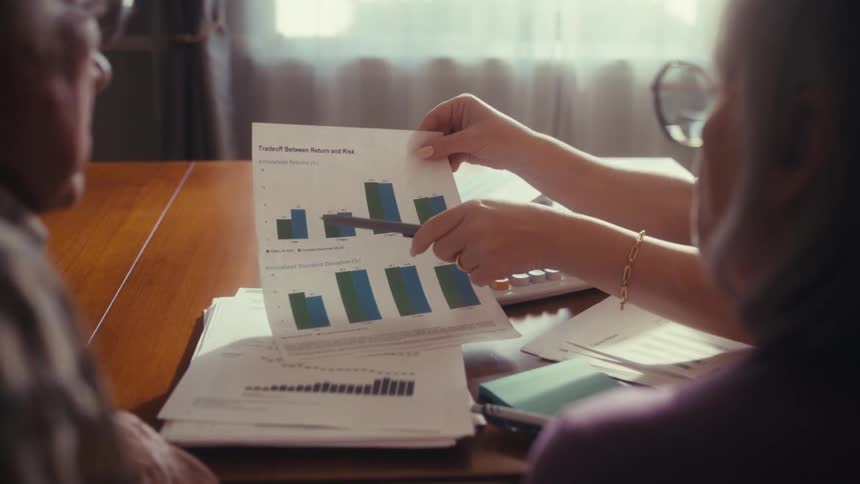 An elderly couple sits at a table while a financial advisor explains their investment performance by pointing to a bar chart