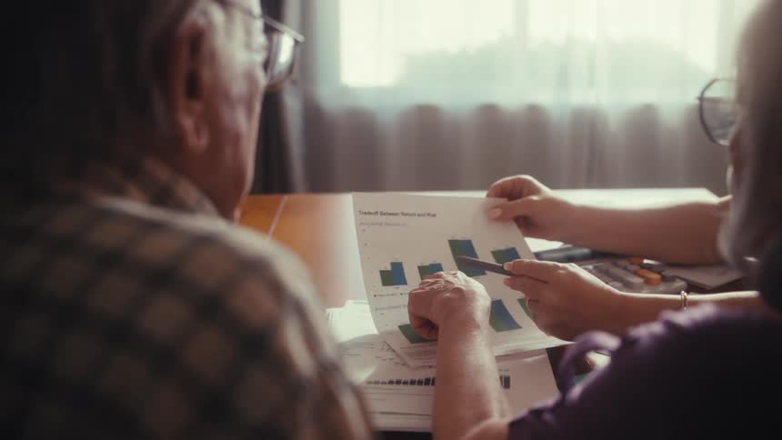 An elderly couple sits at their dining room table reviewing a financial chart together while planning for retirement