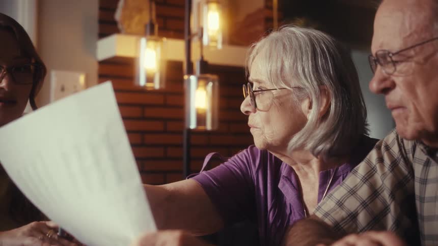 A female consultant explains financial documents to a concerned elderly couple in their home