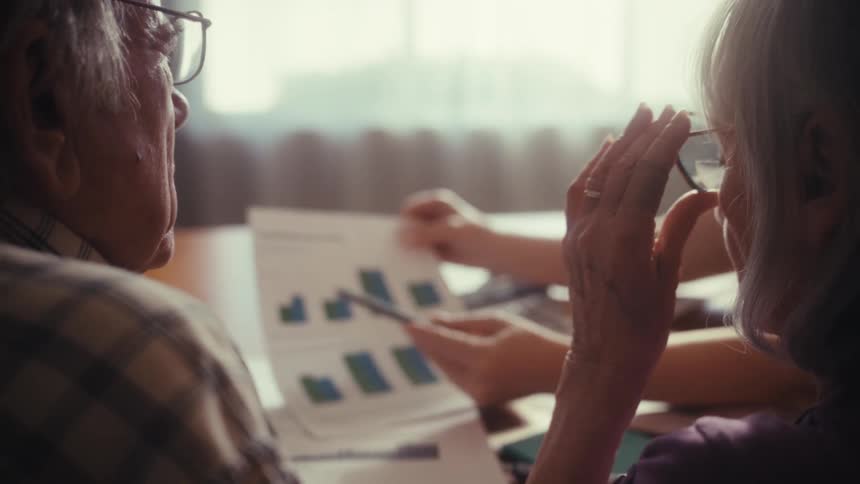 An over-the-shoulder view of a financial advisor explaining bar graphs on a document to an elderly couple in their home