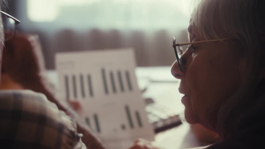 An elderly couple with glasses carefully reviews a financial document with bar graphs, discussing their savings and retirement plan