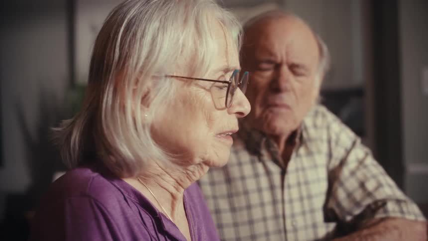 A distressed elderly woman talks with shock and fear while her husband sits worriedly behind her
