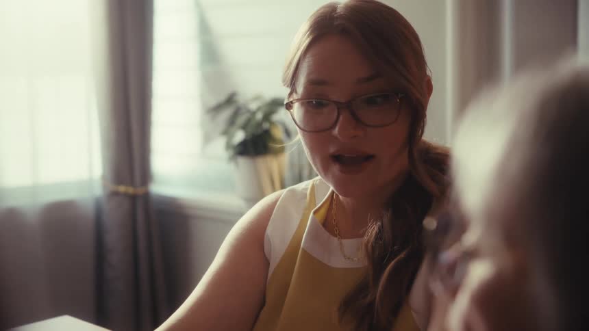 A female financial advisor with glasses explains investment return and risk charts to a senior client during a meeting at home