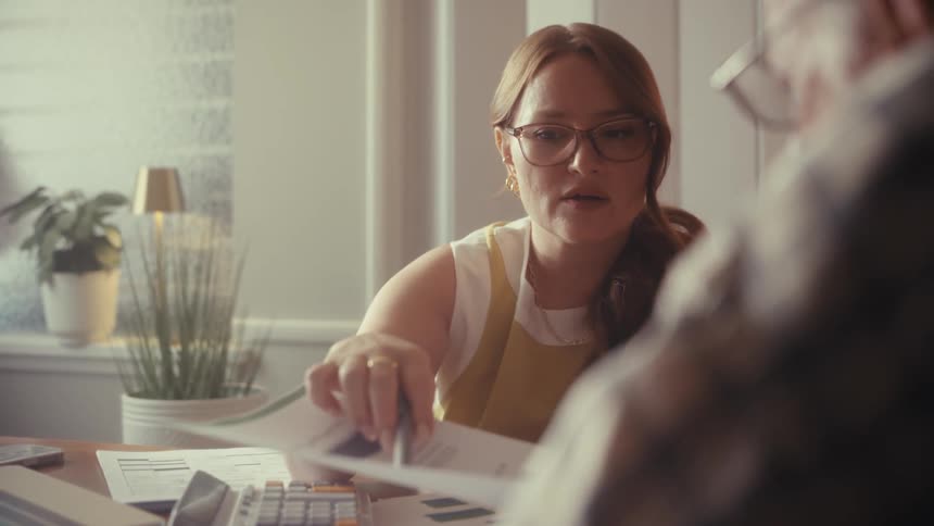 A young businesswoman wearing glasses consults with a senior man, pointing to a financial chart with a pen in a sunlit office