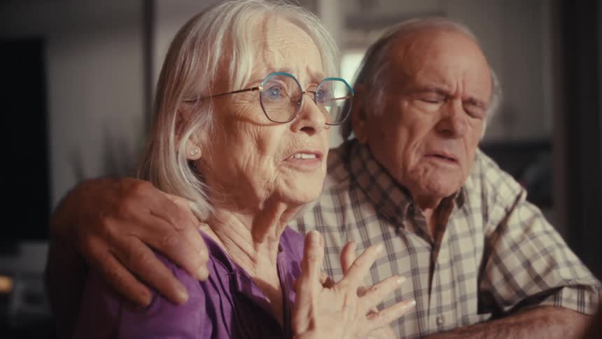 A distraught elderly woman wearing glasses explains a terrible situation as her concerned husband comforts her