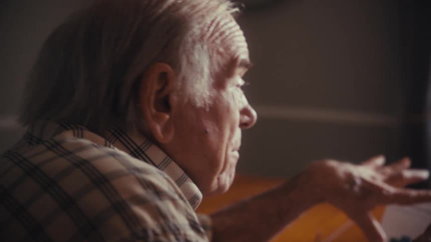 An elderly man gestures while discussing financial documents with his wife and a consultant at a kitchen table