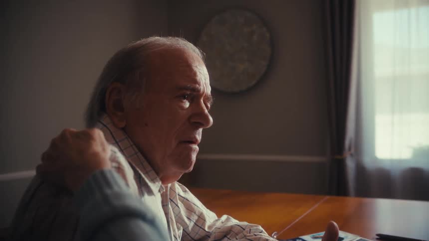 A senior woman with glasses comforts her pensive elderly husband, gently touching his shoulder as they talk in their home