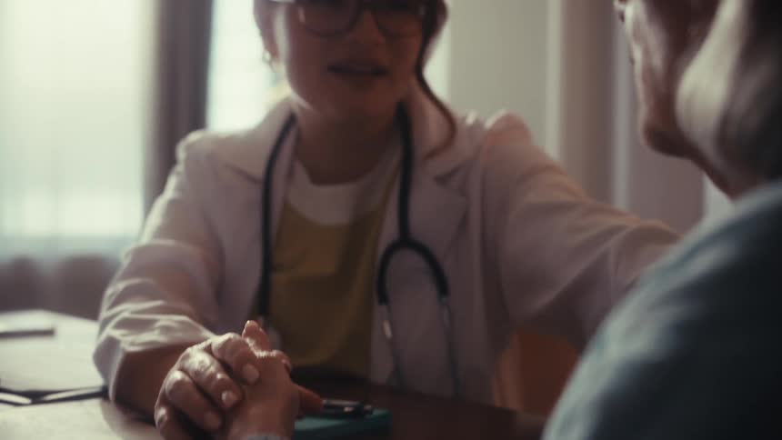 A compassionate young female doctor in a white coat holds an elderly patient's hand to provide comfort and support during a medical consultation