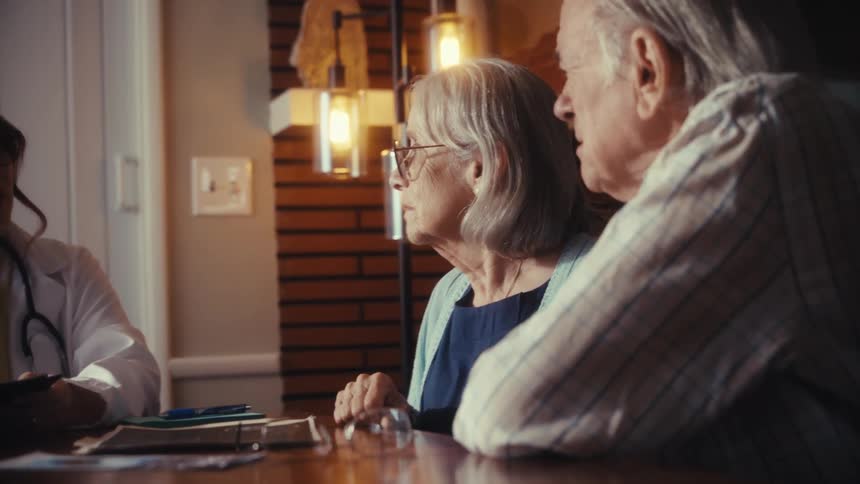 A concerned elderly couple sits at their dining table attentively listening to a female doctor during a home consultation