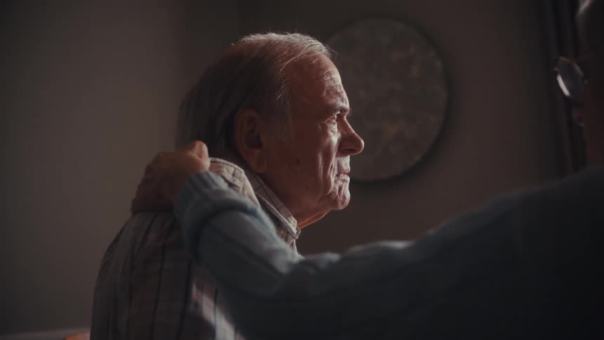 A supportive elderly woman comforts her husband as a doctor delivers somber news during a home visit