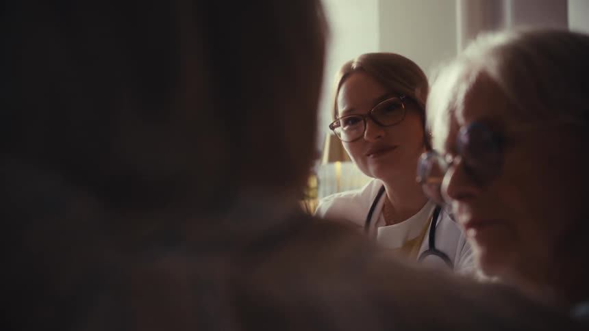 A compassionate female doctor attentively listens to an elderly couple during a serious medical consultation at home