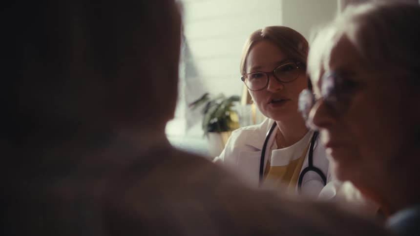 A caring female doctor consults with an elderly couple in a warmly lit room, with the focus shifting to the senior woman listening intently