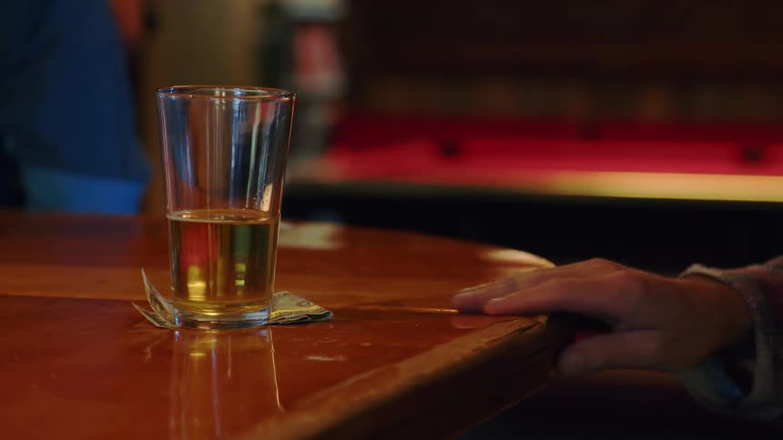 An older man in a plaid shirt takes a sip from a glass of beer at a bar, then leaves it with a dollar bill and walks away