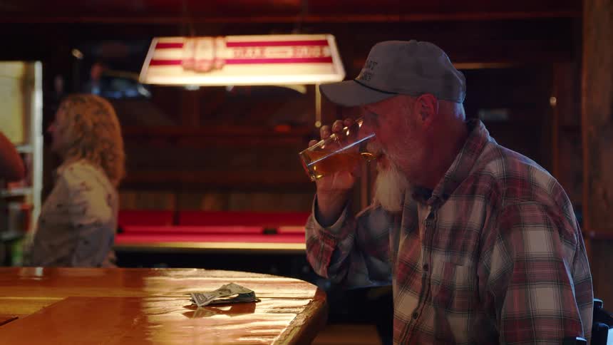 An older man with a white beard and plaid shirt enjoys a beer at a rustic bar before leaving cash on the counter and walking out of frame