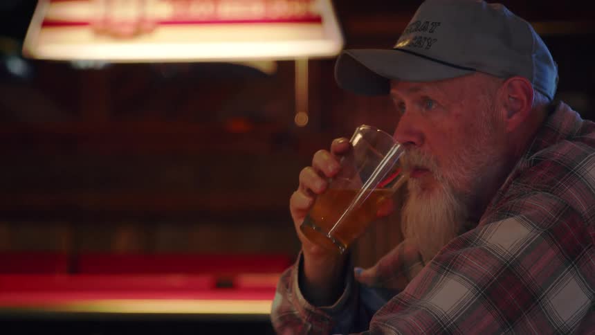 A pensive older man with a long white beard sits alone at a wooden bar counter, slowly sipping a glass of beer