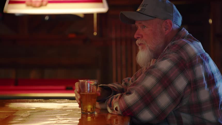 An elderly man with a long white beard sits alone at a rustic bar, thoughtfully drinking a glass of beer