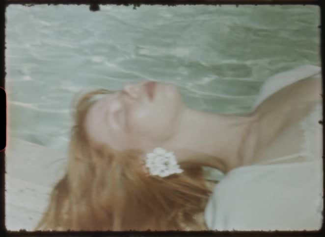A serene red-haired woman with her eyes closed lies peacefully by the rippling water, captured on vintage film