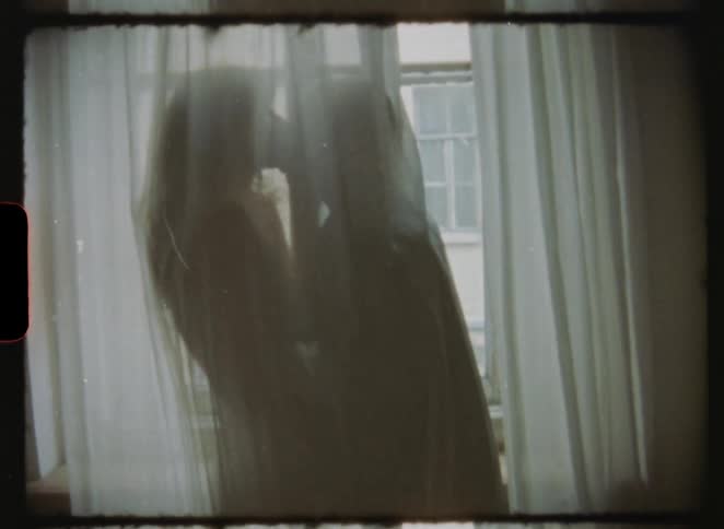 A vintage 8mm film shows the intimate silhouette of a romantic couple kissing behind sheer curtains in front of a window