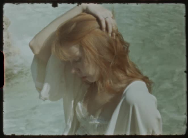 A beautiful red-haired woman in a sheer white robe stands pensively in sunlit water, captured on vintage film