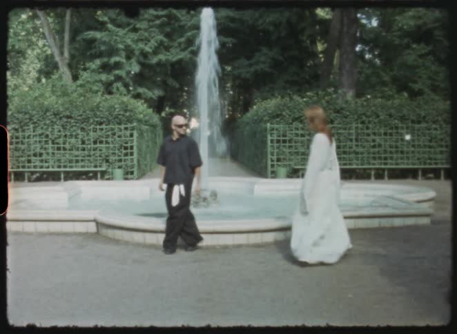 Archival footage captures a man and woman in contrasting outfits walking past a beautiful fountain in a lush green park