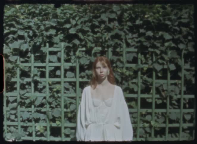 Vintage 8mm footage shows a beautiful red-haired woman in a white robe walking and posing in a sunlit garden against a green ivy trellis