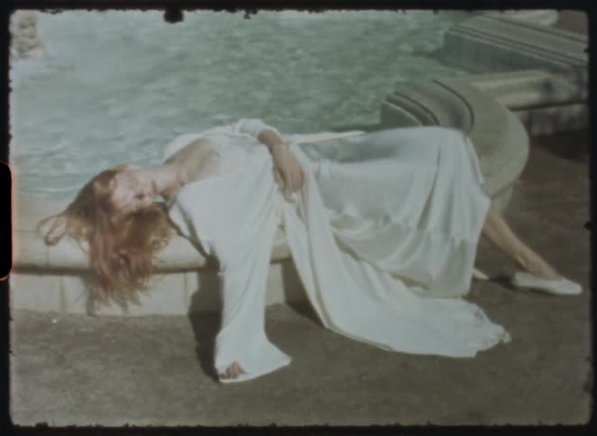 Vintage 8mm footage shows a red-haired woman in a white dress lying languidly by a sunlit fountain