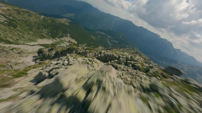 An exhilarating FPV drone flies backward over a rocky mountain ridge, showcasing the vast mountain valley below