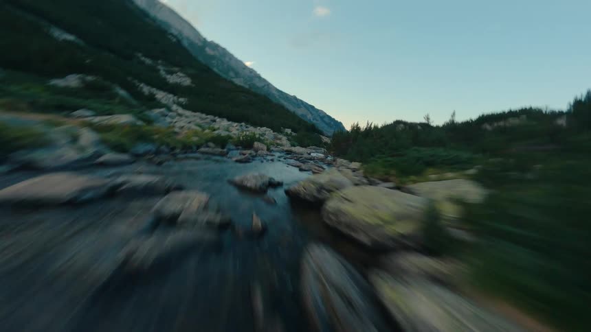 An FPV drone flies low and fast upstream through a rocky mountain valley during the golden hour