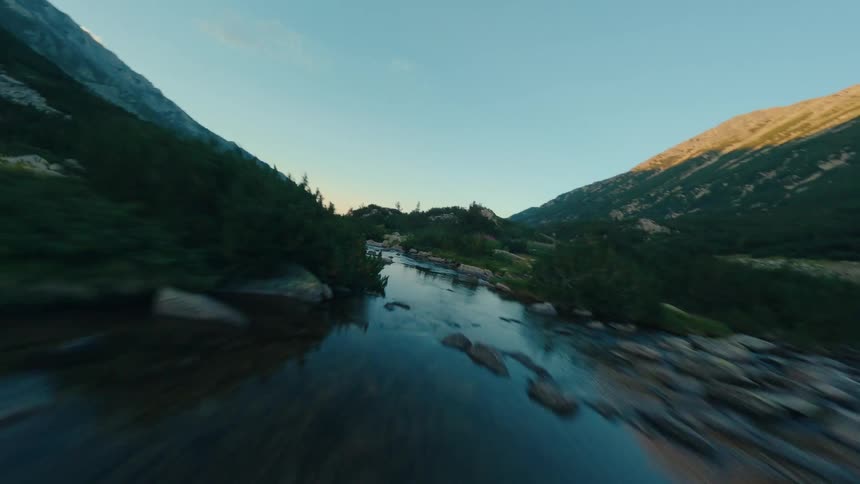 A dynamic FPV drone shot soars backward over a serene mountain stream nestled between majestic peaks at dawn