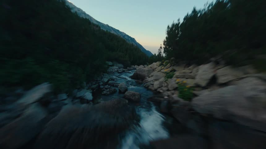 A dynamic FPV drone flies backward at high speed over a rushing mountain river in a rocky canyon at dusk