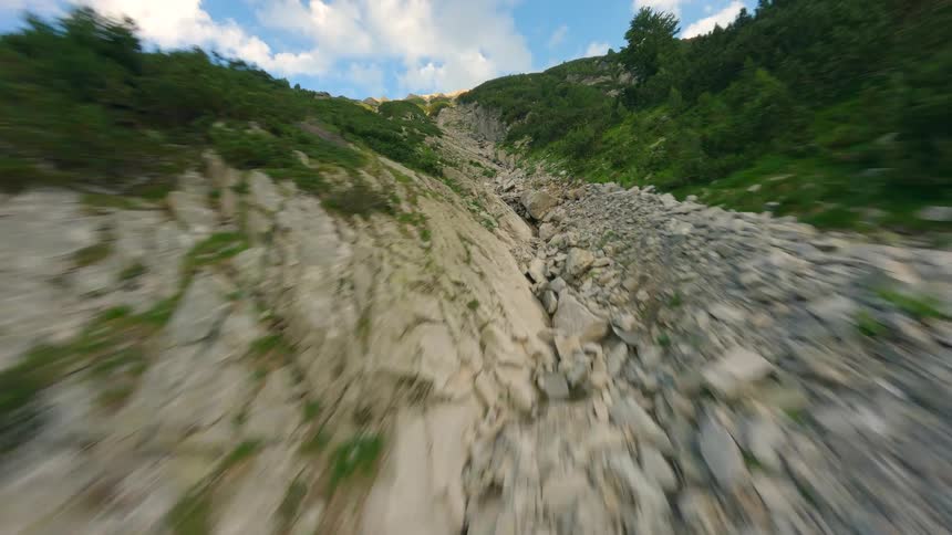 A fast-paced FPV drone flight soaring up a rugged, stony ravine on a green mountainside