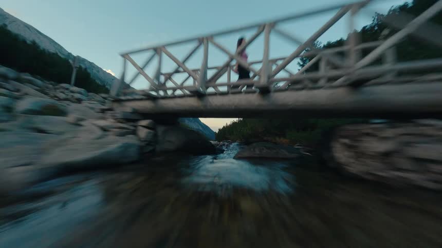 A thrilling FPV drone shot follows a rocky river upstream through a mountain valley, passing under a wooden bridge