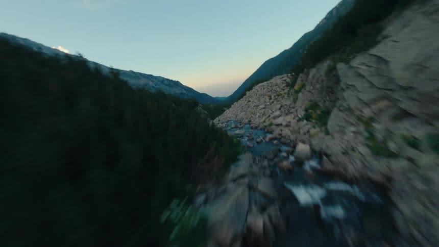 An exhilarating FPV drone shot races up a rocky mountain river, ascending to reveal a vast, lush green valley at dusk