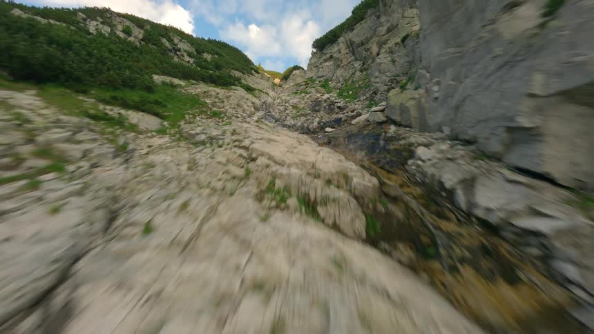 A dizzying FPV drone flight backwards down a steep, rocky mountain slope with patches of green vegetation