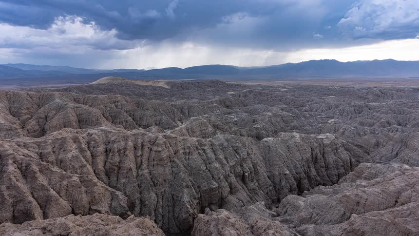 A dramatic timelapse captures a powerful storm cell releasing rain over a vast and rugged desert badlands landscape