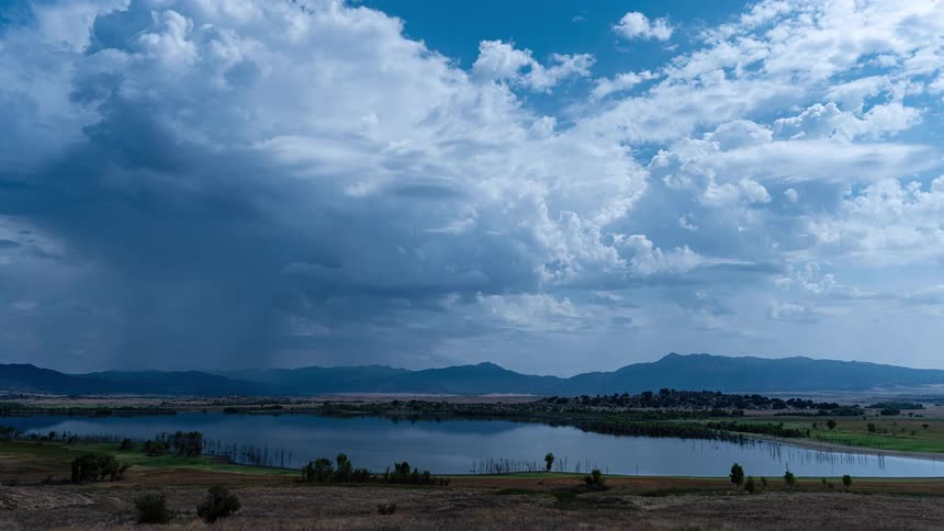 Ominous storm clouds and falling rain drift across the sky above a tranquil mountain lake in a dramatic cinemagraph