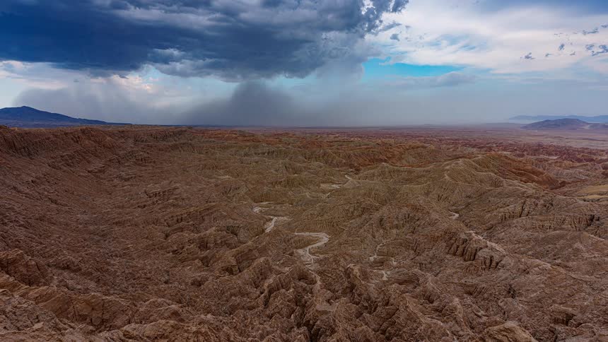 An epic timelapse captures a localized rainstorm moving over the rugged and expansive desert badlands under dramatic clouds