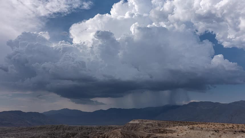 A powerful storm cloud releases a downpour of rain over a vast and arid desert mountain landscape