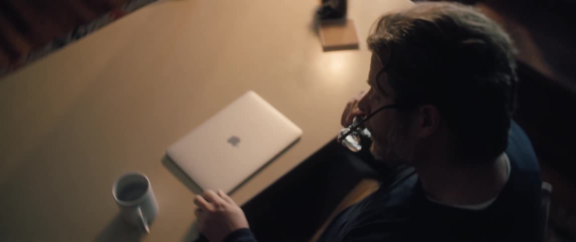 A thoughtful man takes off his glasses while sitting at a table with his closed laptop and a mug in a dimly lit room