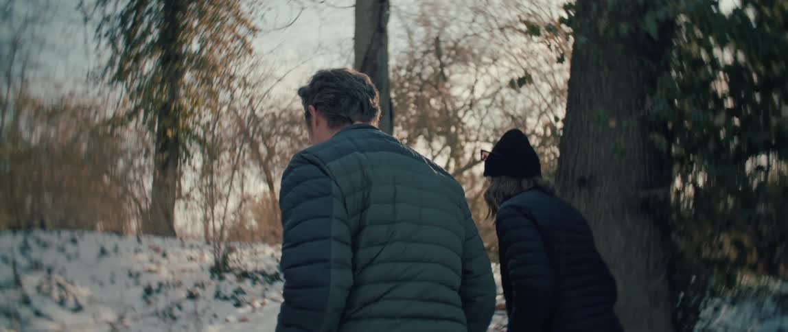 A couple wearing winter puffer jackets walk through a snowy forest on a sunny day, seen from a low angle from behind