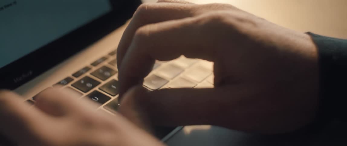 A close-up slow-motion shot of a man's hands with a wedding ring typing on a laptop keyboard in a dimly lit room