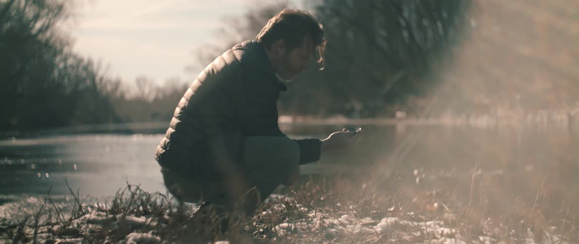 A man in a winter coat thoughtfully examines a small found object while kneeling by a sunlit, frozen river in slow motion