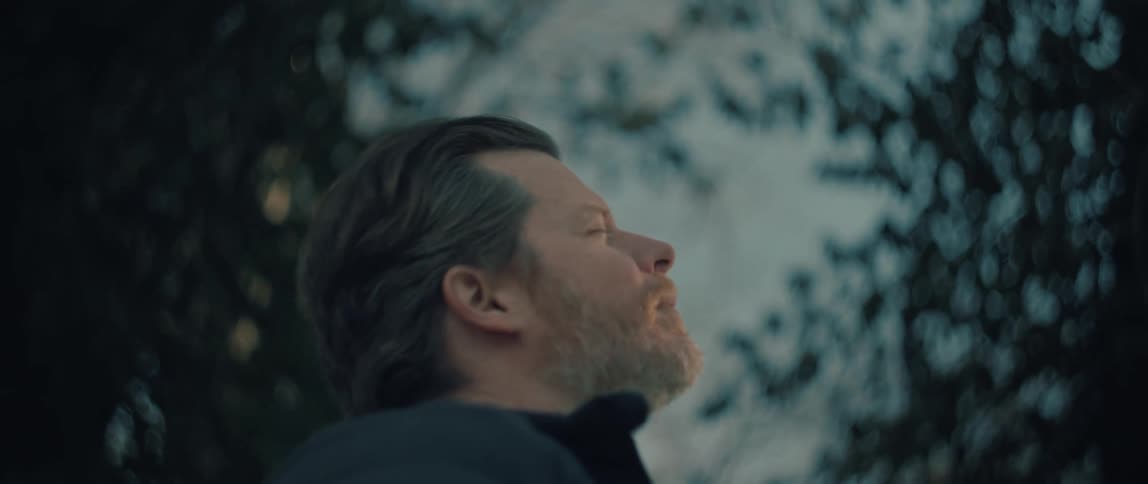 A serene man with a beard closes his eyes and breathes in the cold, crisp air, his face warmed by the gentle sunlight