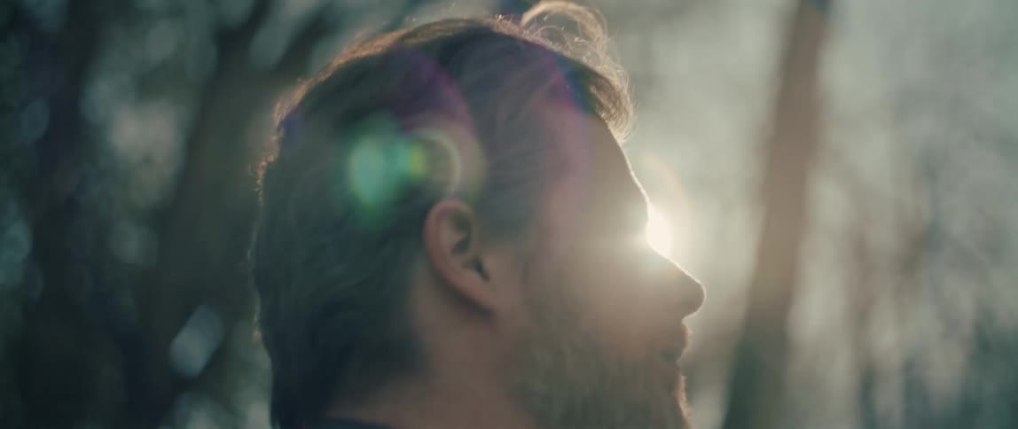 A contemplative man with a beard enjoys the golden hour sunlight in a forest before turning and walking away in slow motion