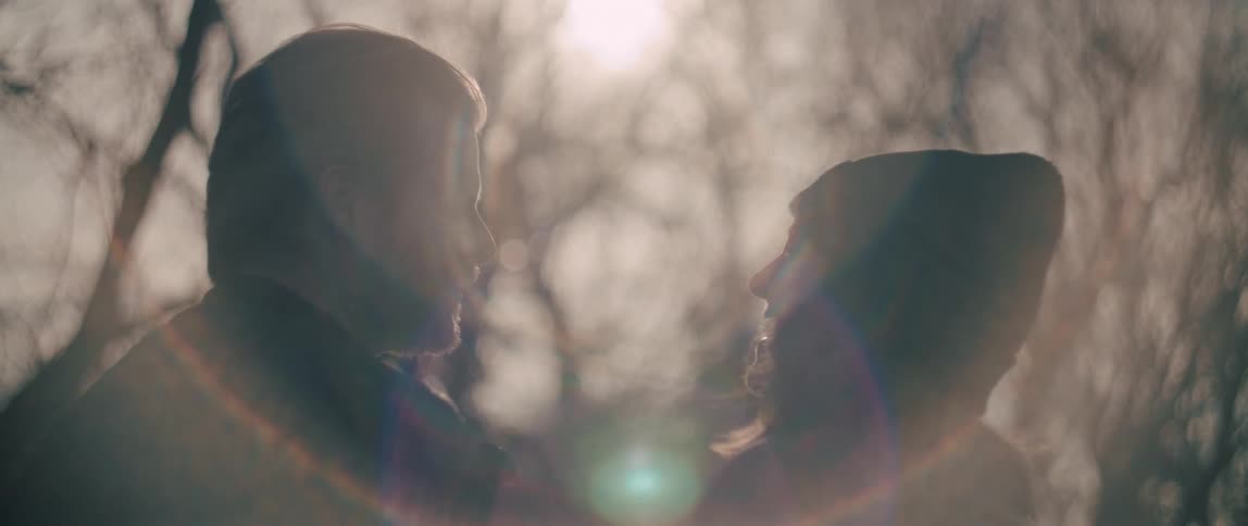 A slow focus pull reveals the soft silhouettes of a man and woman sharing an intimate moment in a sun-flared forest