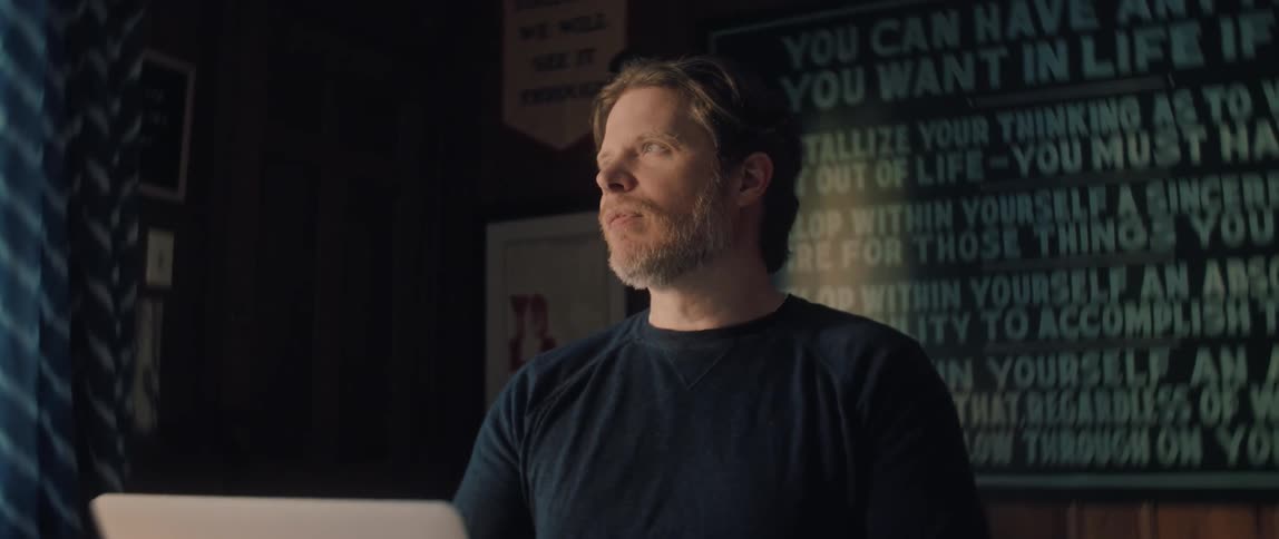 A focused man with a beard looks up from his laptop while working in a cozy, dark room