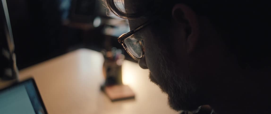 A focused man with a beard puts on his glasses in slow motion while working at his desk in a dimly lit office