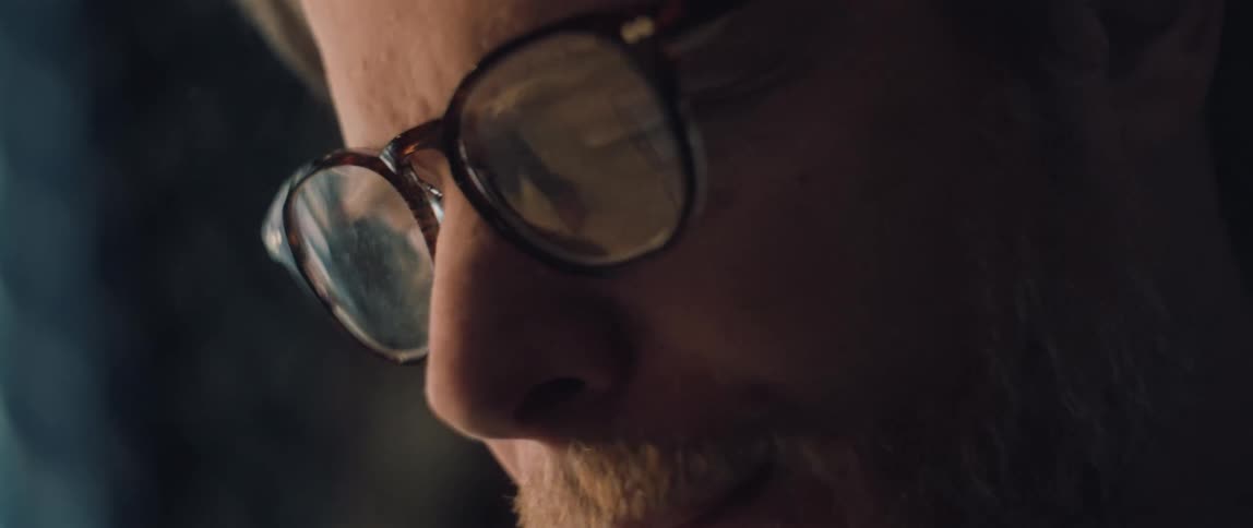 A close-up of a bearded man's glasses reflecting the digital screen he is intently focused on
